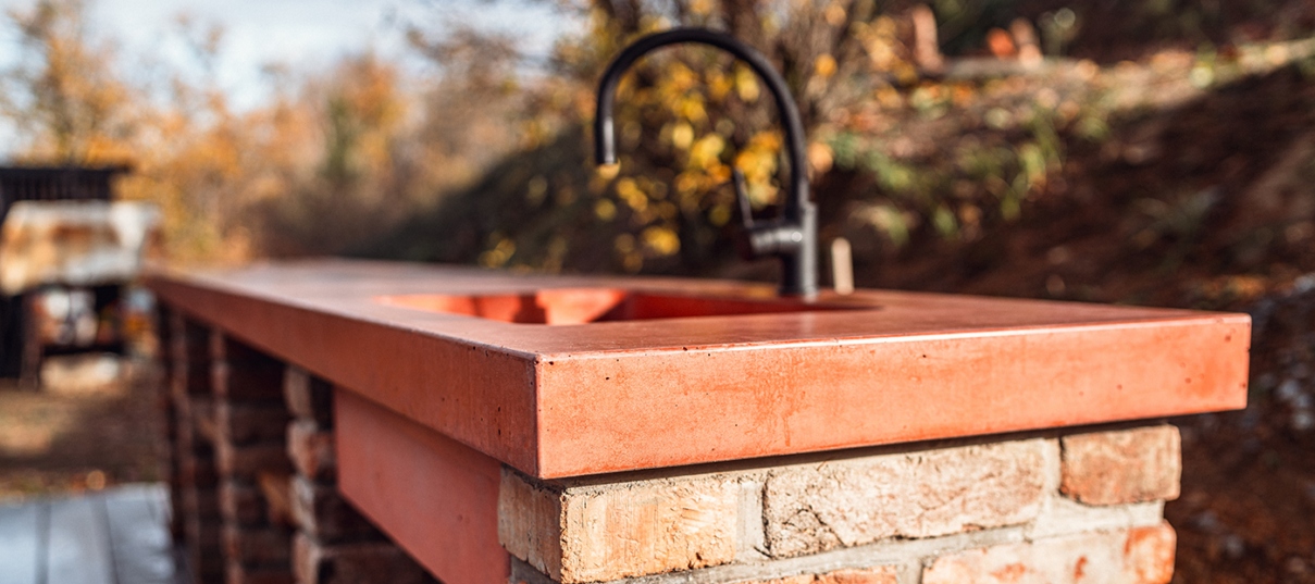 Betonové zahradní vybavení sloužící jako zahradní kuchyňská linka s pracovní deskou a dřezem z červeného betonu.